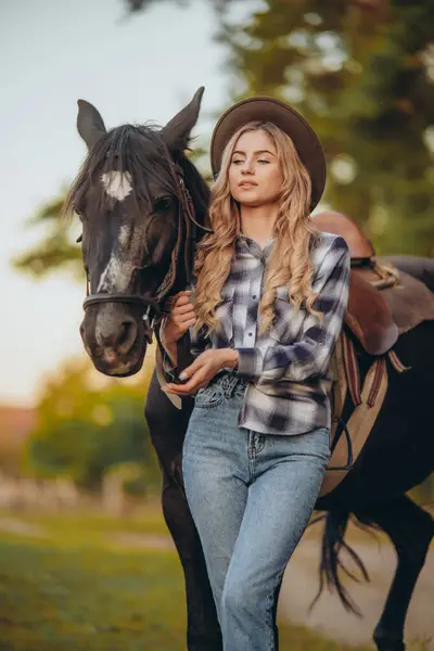 stock image A young woman with a horse stands near the fence on her ranch at sunset in the fall. Breeding thoroughbred horses for equestrian sport.