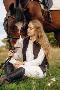 Young beautiful female jockey sits on a meadow near her horse at sunset. Walk with a horse in the summer on a meadow.