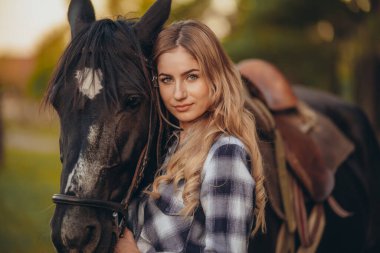 A young woman with a horse stands near the fence on her ranch at sunset in the fall. Breeding thoroughbred horses for equestrian sport. clipart