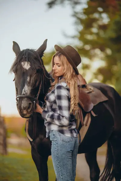 stock image A young woman with a horse stands near the fence on her ranch at sunset in the fall. Breeding thoroughbred horses for equestrian sport.