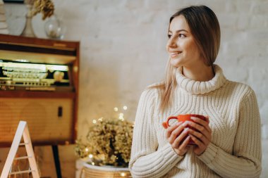 Genç ve güzel bir kadın, Noel arifesinde pencerenin yanındaki bir evde elinde bir kupa sıcak çayla oturuyor..