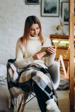 Genç, güzel bir kadın, battaniyeyle kaplı bir sandalyede oturur ve Noel arifesinde pencerenin yanında kitap okur. Noel arifesinde rahat bir sandalyede kitap okumak..