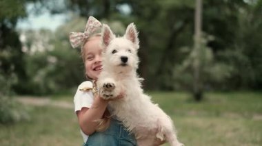 Sevimli küçük bir kız, parkta yürürken en sevdiği küçük Batı Highland Beyaz Teriyer köpeğini kollarında tutuyor..