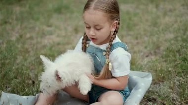 Sevimli küçük bir kız, parkta yürürken en sevdiği küçük Batı Highland Beyaz Teriyer köpeğini kollarında tutuyor..