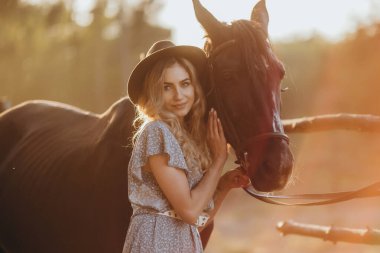 Atı olan genç bir kadın sonbaharda günbatımında çiftliğinde çitin yanında duruyor. Binicilik sporu için safkan atlar yetiştiriyor..