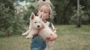 Sevimli küçük bir kız, parkta yürürken en sevdiği küçük Batı Highland Beyaz Teriyer köpeğini kollarında tutuyor..