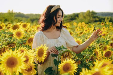 Genç, tatlı esmer kız yazın gün batımında çiçek açan ayçiçeği tarlasında duruyor. Ayçiçeği yağı üretiminde artış.