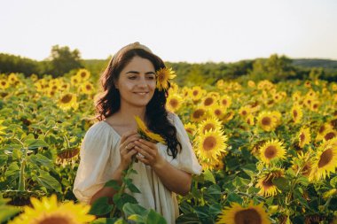 Genç, tatlı esmer kız yazın gün batımında çiçek açan ayçiçeği tarlasında duruyor. Ayçiçeği yağı üretiminde artış.