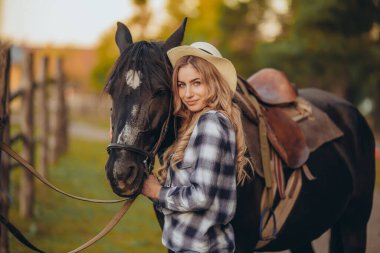 Atı olan genç bir kadın sonbaharda günbatımında çiftliğinde çitin yanında duruyor. Binicilik sporu için safkan atlar yetiştiriyor..