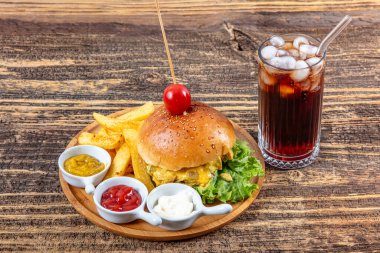 Eski ahşap bir masada et, peynir ve sebzeli nefis ev yapımı hamburger. Patates kızartması, ketçap ve mayonez ile servis edilen buzlu soğuk içecek..