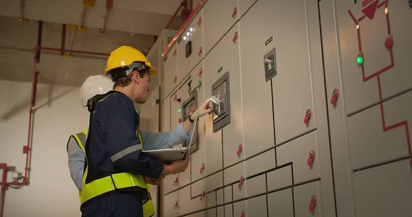 Two electricians electrical engineers in protective uniform checking voltage control panel screen system at electrical cabinet for generate electricity of factory in manufacture industrial