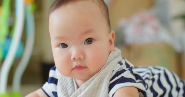 Meraklı, masum Asyalı, koca gözlü, çizgili elbiseli ve yumuşak önlüklü bir bebek. Evdeki oyuncakların bulanık arka planıyla kısmen etrafa bakıyor.