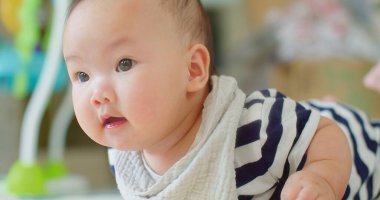 Meraklı, masum Asyalı, koca gözlü, çizgili elbiseli ve yumuşak önlüklü bir bebek. Evdeki oyuncakların bulanık arka planıyla kısmen etrafa bakıyor.