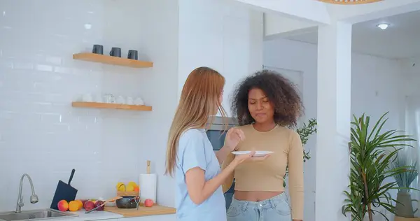 stock image Laughter fills a sunlit kitchen as one woman presents a plate of food to her friend, who reacts with a delighted surprise