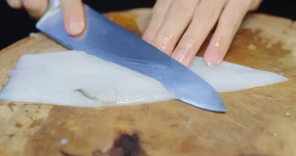 Detailed view of a hand slicing through white translucent squid on a well-worn wooden cutting board, showcasing seafood preparation skills technique