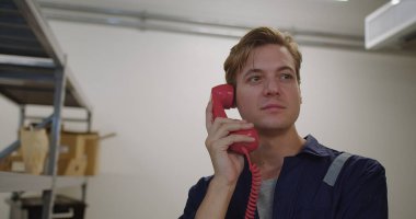 İş üniformalı endişeli bir adam depoda kırmızı bir telefonla konuşuyor.