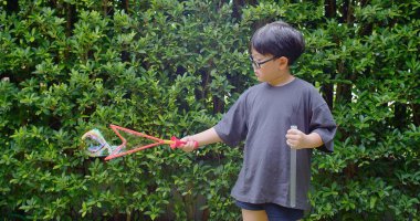 Mutlu, genç Asyalı çocuk gülümseyerek baloncuklu sabun oyuncağı havada sallanırken arka bahçedeki yeşil bahçeye karşı yavaş çekimde, yaz tatili eğlencesi çocukluğunda eğleniyor.