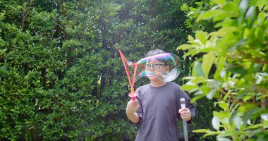 Mutlu, genç Asyalı çocuk gülümseyerek baloncuklu sabun oyuncağı havada sallanırken arka bahçedeki yeşil bahçeye karşı yavaş çekimde, yaz tatili eğlencesi çocukluğunda eğleniyor.