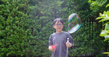 Mutlu, genç Asyalı çocuk gülümseyerek baloncuklu sabun oyuncağı havada sallanırken arka bahçedeki yeşil bahçeye karşı yavaş çekimde, yaz tatili eğlencesi çocukluğunda eğleniyor.