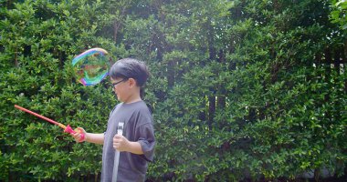 Mutlu, genç Asyalı çocuk gülümseyerek baloncuklu sabun oyuncağı havada sallanırken arka bahçedeki yeşil bahçeye karşı yavaş çekimde, yaz tatili eğlencesi çocukluğunda eğleniyor.