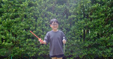 Mutlu, genç Asyalı çocuk gülümseyerek baloncuklu sabun oyuncağı havada sallanırken arka bahçedeki yeşil bahçeye karşı yavaş çekimde, yaz tatili eğlencesi çocukluğunda eğleniyor.