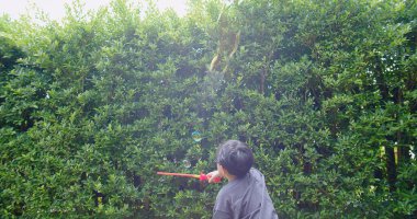 Mutlu, genç Asyalı çocuk gülümseyerek baloncuklu sabun oyuncağı havada sallanırken arka bahçedeki yeşil bahçeye karşı yavaş çekimde, yaz tatili eğlencesi çocukluğunda eğleniyor.
