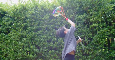 Mutlu, genç Asyalı çocuk gülümseyerek baloncuklu sabun oyuncağı havada sallanırken arka bahçedeki yeşil bahçeye karşı yavaş çekimde, yaz tatili eğlencesi çocukluğunda eğleniyor.