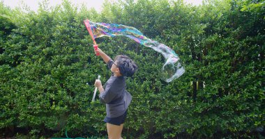 Mutlu, genç Asyalı çocuk gülümseyerek baloncuklu sabun oyuncağı havada sallanırken arka bahçedeki yeşil bahçeye karşı yavaş çekimde, yaz tatili eğlencesi çocukluğunda eğleniyor.