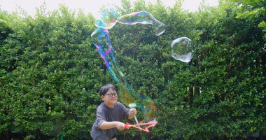 Mutlu, genç Asyalı çocuk gülümseyerek baloncuklu sabun oyuncağı havada sallanırken arka bahçedeki yeşil bahçeye karşı yavaş çekimde, yaz tatili eğlencesi çocukluğunda eğleniyor.