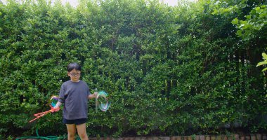 Mutlu, genç Asyalı çocuk gülümseyerek baloncuklu sabun oyuncağı havada sallanırken arka bahçedeki yeşil bahçeye karşı yavaş çekimde, yaz tatili eğlencesi çocukluğunda eğleniyor.