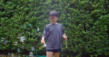 Mutlu, genç Asyalı çocuk gülümseyerek baloncuklu sabun oyuncağı havada sallanırken arka bahçedeki yeşil bahçeye karşı yavaş çekimde, yaz tatili eğlencesi çocukluğunda eğleniyor.