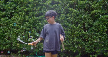 Mutlu, genç Asyalı çocuk gülümseyerek baloncuklu sabun oyuncağı havada sallanırken arka bahçedeki yeşil bahçeye karşı yavaş çekimde, yaz tatili eğlencesi çocukluğunda eğleniyor.