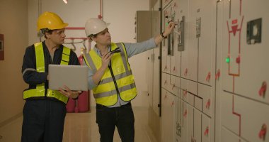 İki elektrikçi koruyucu üniformalı elektrik mühendisi elektrik kabinindeki voltaj kontrol paneli sistemini kontrol ederek endüstriyel üretim fabrikasında elektrik üretiyor.