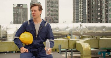 Portre yakışıklı, yorgun, Kafkasyalı profesyonel inşaat mühendisi üniformalı ve sert şapkalı mühendis dinleniyor. Çatı katındaki şantiyede mola verip su içiyor.