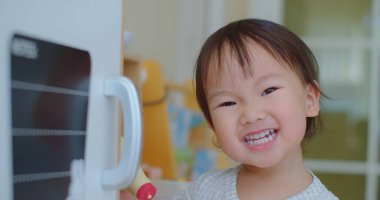 Neşeli bebek ışıltılı bir gülümseme ile, beyaz bir fırının yanında duruyor parlak bir oyuncak mutfak ortamında