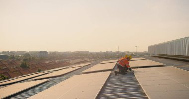 Güneş enerjisi teknisyenleri yansıtıcı teçhizatlarla güneş tesislerini gün batımında, banliyö arkaplanıyla birlikte çatı panellerinde incelemeyi tartışıyorlar.