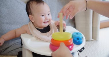 Neşeli bebek sandalyeye oturur ve annesinin yardımıyla renkli yüzükler takmayı öğrenir. Çocukluk gelişimi aktiviteleriyle meşgul olan bir an..