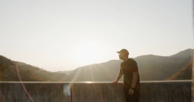 Bir adam bir dağ sırasının tepesine bakıyor. Güneş ışıl ışıl parlıyor, sahne üzerine sıcak bir parıltı saçıyor. Adam manzaranın tadını çıkarıyor ve manzaranın güzelliğini içine çekiyor.