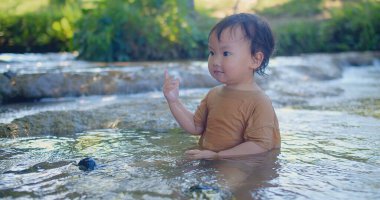 Coşkulu çocuk sığ bir dereye sıçrıyor, güneş ışığı gibi parıldıyor yeşil yaprakların arasından mutluluk dolu bir açık hava sahnesinde.