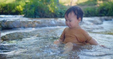 Coşkulu çocuk sığ bir dereye sıçrıyor, güneş ışığı gibi parıldıyor yeşil yaprakların arasından mutluluk dolu bir açık hava sahnesinde.