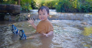 Coşkulu çocuk sığ bir dereye sıçrıyor, güneş ışığı gibi parıldıyor yeşil yaprakların arasından mutluluk dolu bir açık hava sahnesinde.