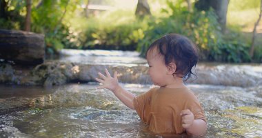 Kahverengi gömlekli meraklı bir çocuk, su sıçratma hissini zevkle keşfeder. Su, yemyeşil bitki örtüsüyle çevrili sığ bir derede oynar.