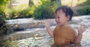 Kahverengi gömlekli meraklı bir çocuk, su sıçratma hissini zevkle keşfeder. Su, yemyeşil bitki örtüsüyle çevrili sığ bir derede oynar.