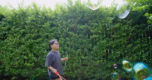 Mutlu, genç Asyalı çocuk gülümseyerek baloncuklu sabun oyuncağı havada sallanırken arka bahçedeki yeşil bahçeye karşı yavaş çekimde, yaz tatili eğlencesi çocukluğunda eğleniyor.