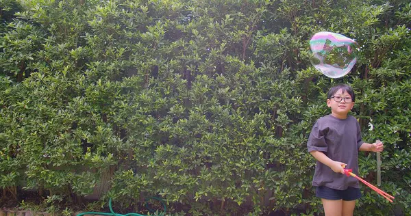 Mutlu, genç Asyalı çocuk gülümseyerek baloncuklu sabun oyuncağı havada sallanırken arka bahçedeki yeşil bahçeye karşı yavaş çekimde, yaz tatili eğlencesi çocukluğunda eğleniyor.