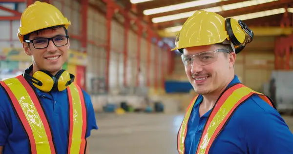 Two happy male industrial workers engineers wearing safety gear hard hats and high visibility vests standing smile confidently in a busy industrial warehouse