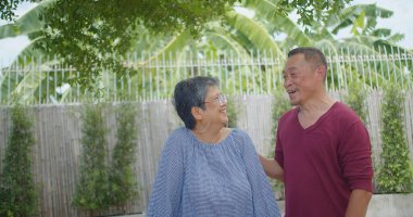 Yaşlı bir emekli Asyalı evli çift güneşli bir bahçede kameraya gülümserek neşeli bir anı paylaşıyor. Sahne mutlu ve rahat.