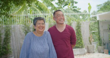 Yaşlı bir emekli Asyalı evli çift güneşli bir bahçede kameraya gülümserek neşeli bir anı paylaşıyor. Sahne mutlu ve rahat.