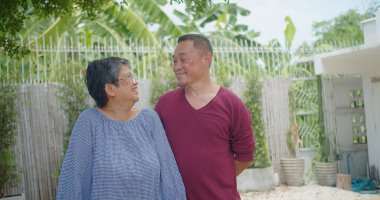 Yaşlı bir emekli Asyalı evli çift güneşli bir bahçede kameraya gülümserek neşeli bir anı paylaşıyor. Sahne mutlu ve rahat.
