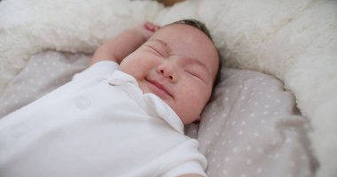 Bir bebek beyaz bir battaniyeyle bir yatakta uyuyor. Bebek gülümsüyor ve huzurlu bir ifadesi var.